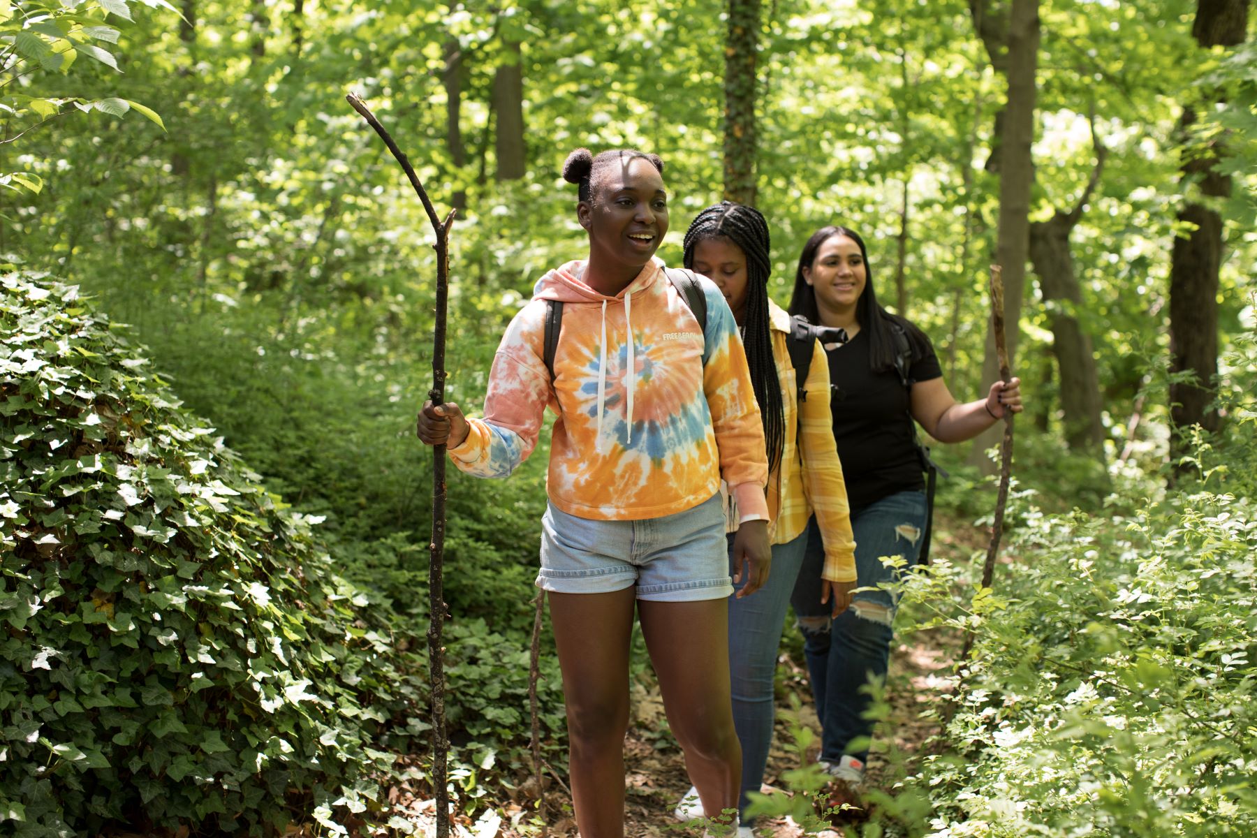 A group of young women with a variety of darker skin tones are hiking along a forest trail and smiling.