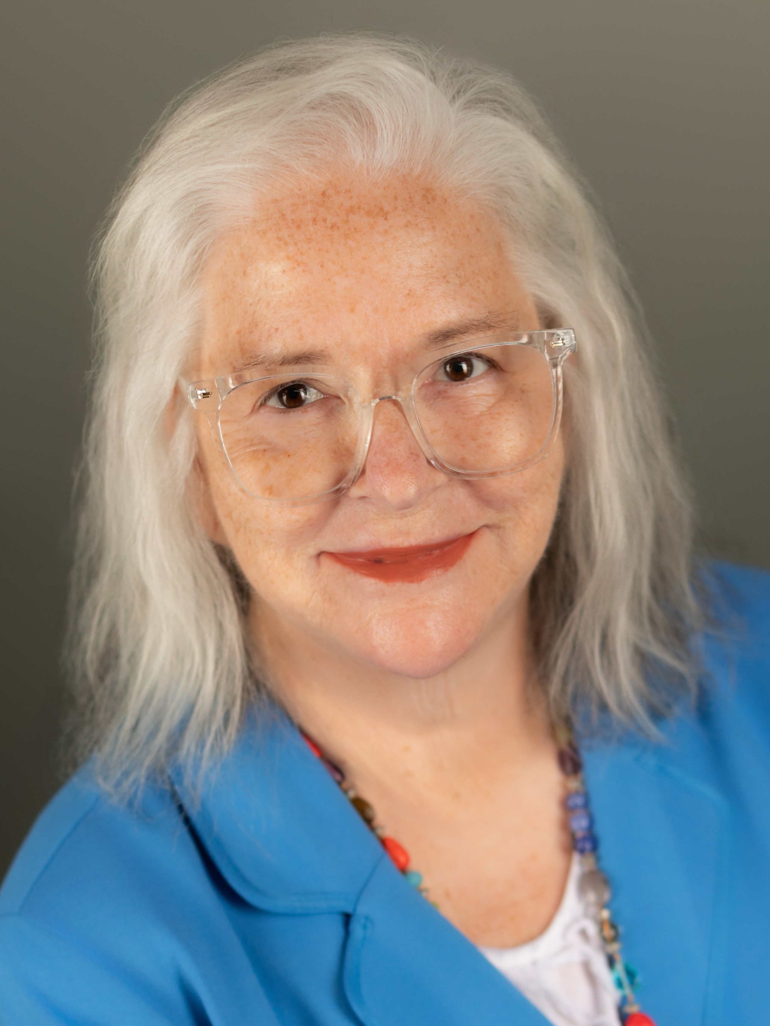 Headshot of Deb Brown, a light skinned woman wearing clear-framed glasses with short white hair wearing a colorful top.