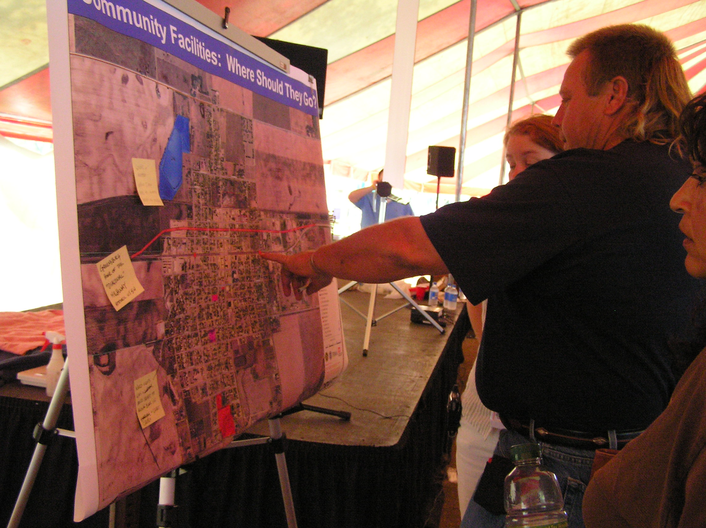 A group of people are looking at a map of a small town. The heading says, "Community Facilities: where should they go?"