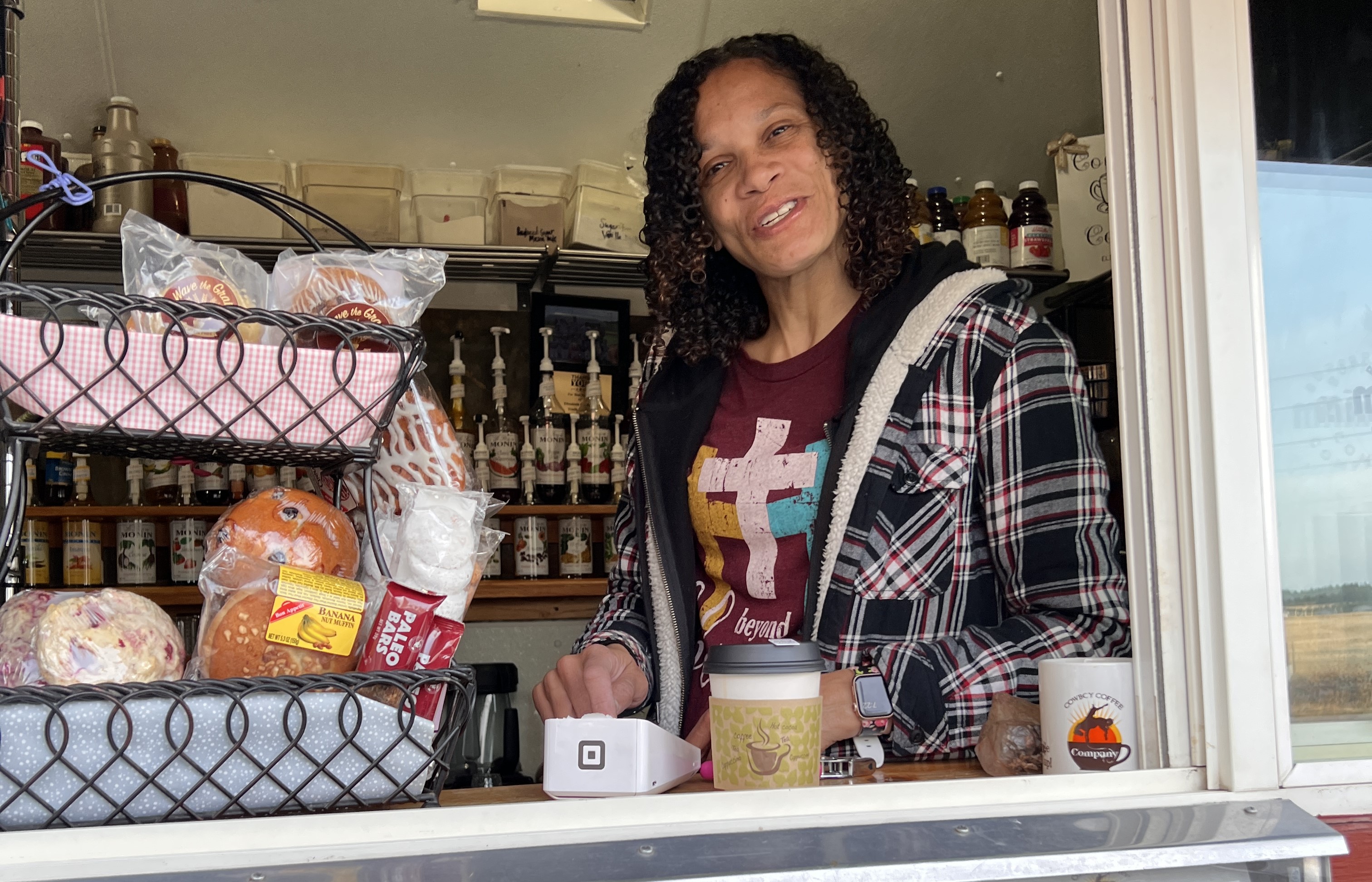 A medium skinned woman with curly hair selling coffee and baked goods from a drive up window.