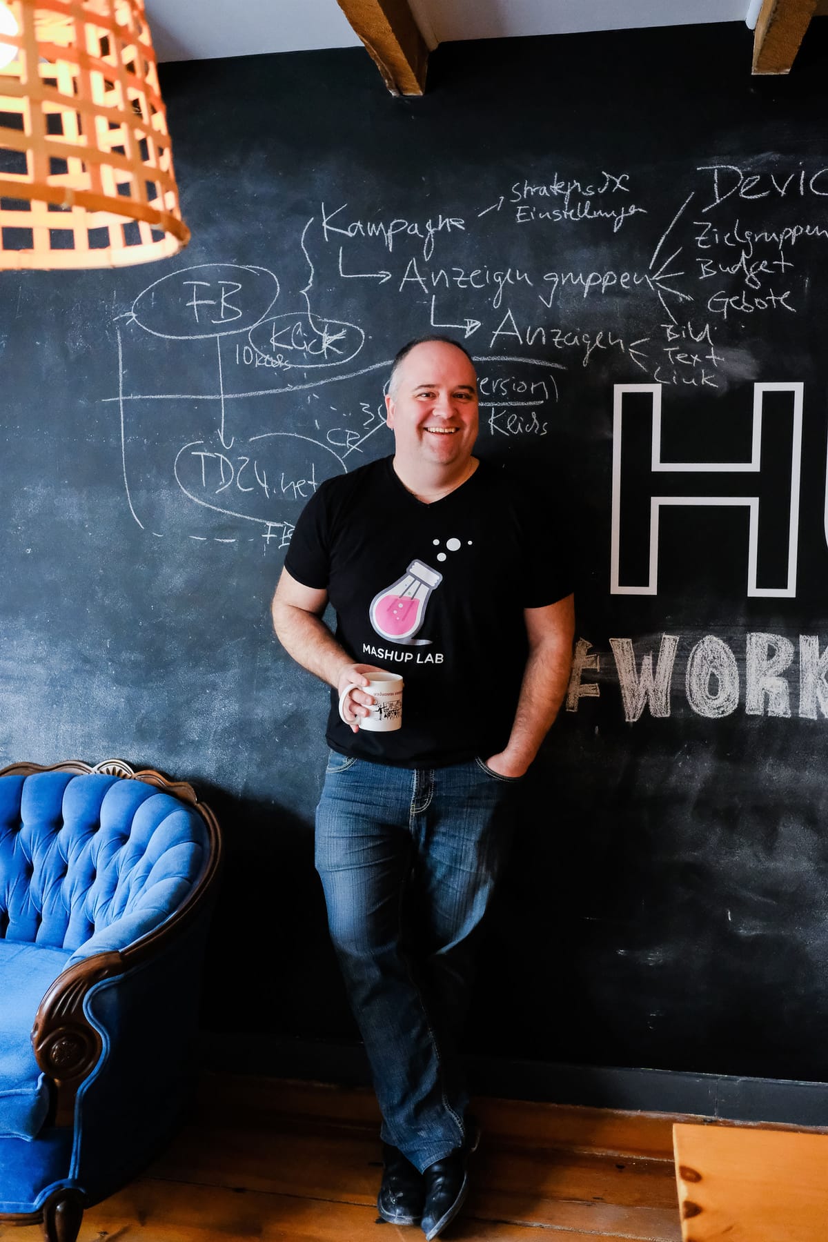 Andrew Button, a man with short hair wearing t-shirt with the Mashup Labs lightbulb logo, is holding a coffee mug and standing in front of a chalkboard