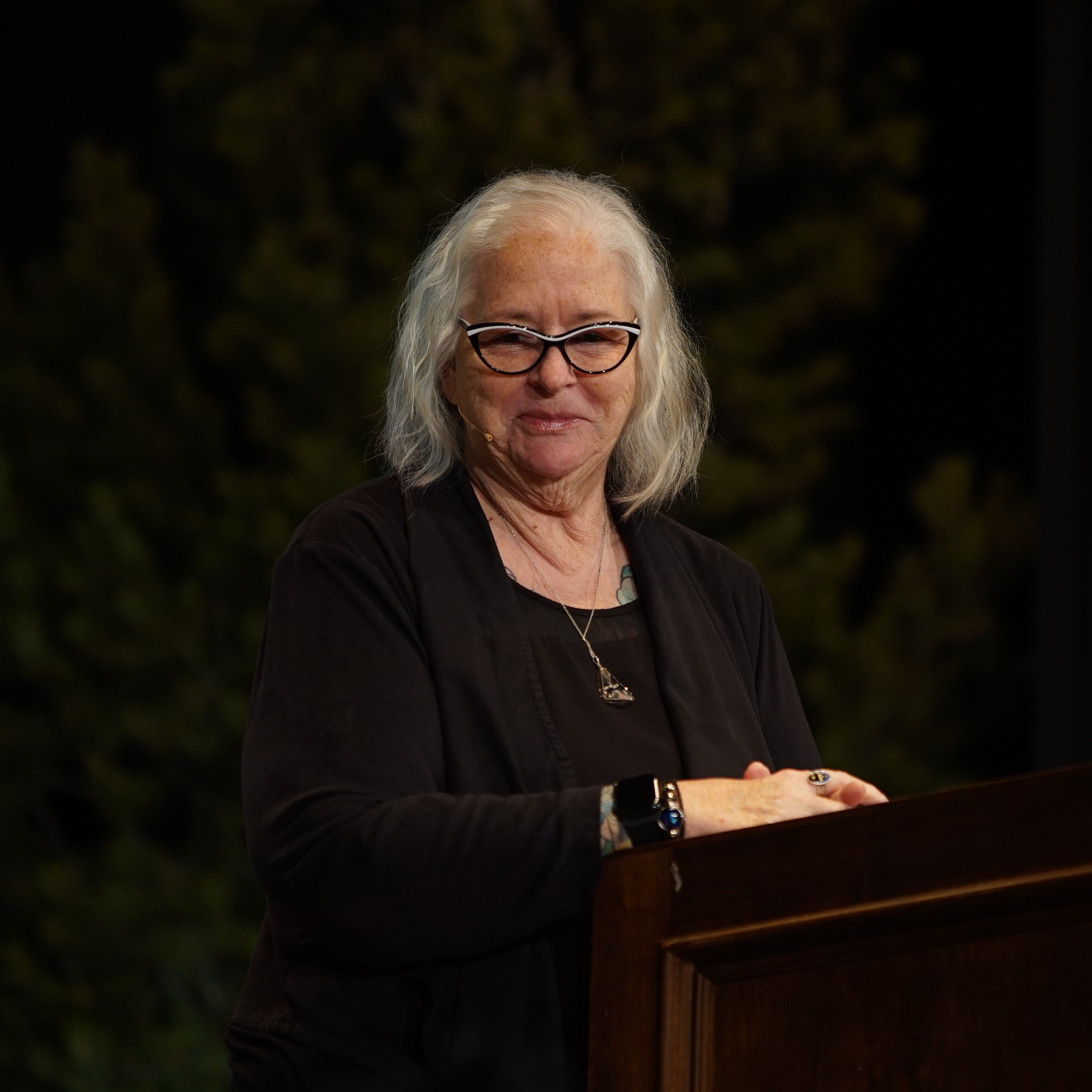 Deb Brown, a light skinned woman wearing cats-eye glasses with mid-length white hair with her hands on a podium.