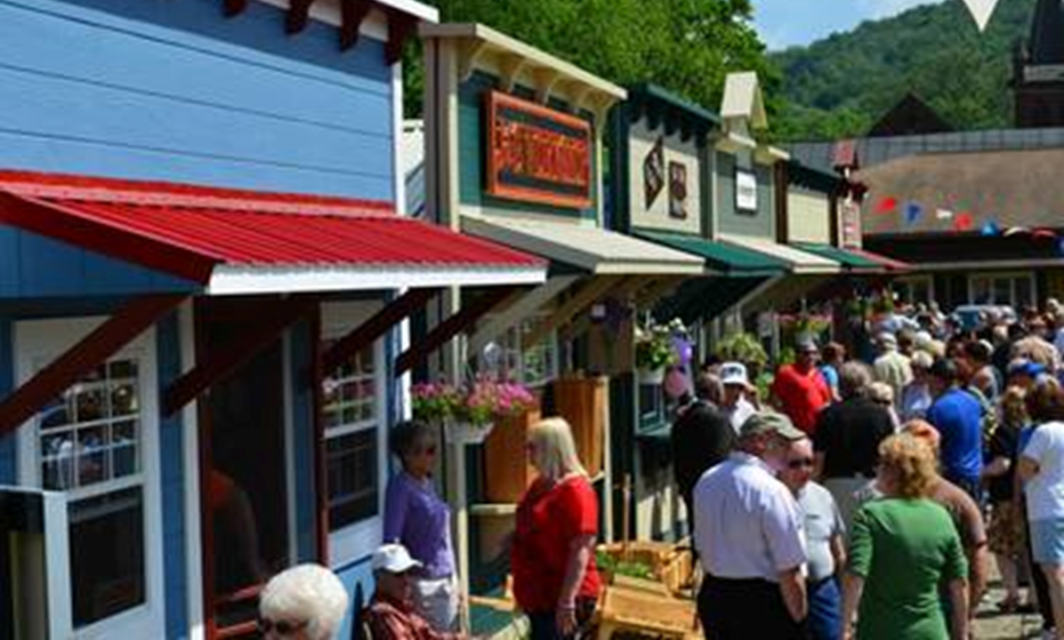 A row of small storage sheds that have been decorated as tiny business storefronts. People are shopping and holding conversations. 