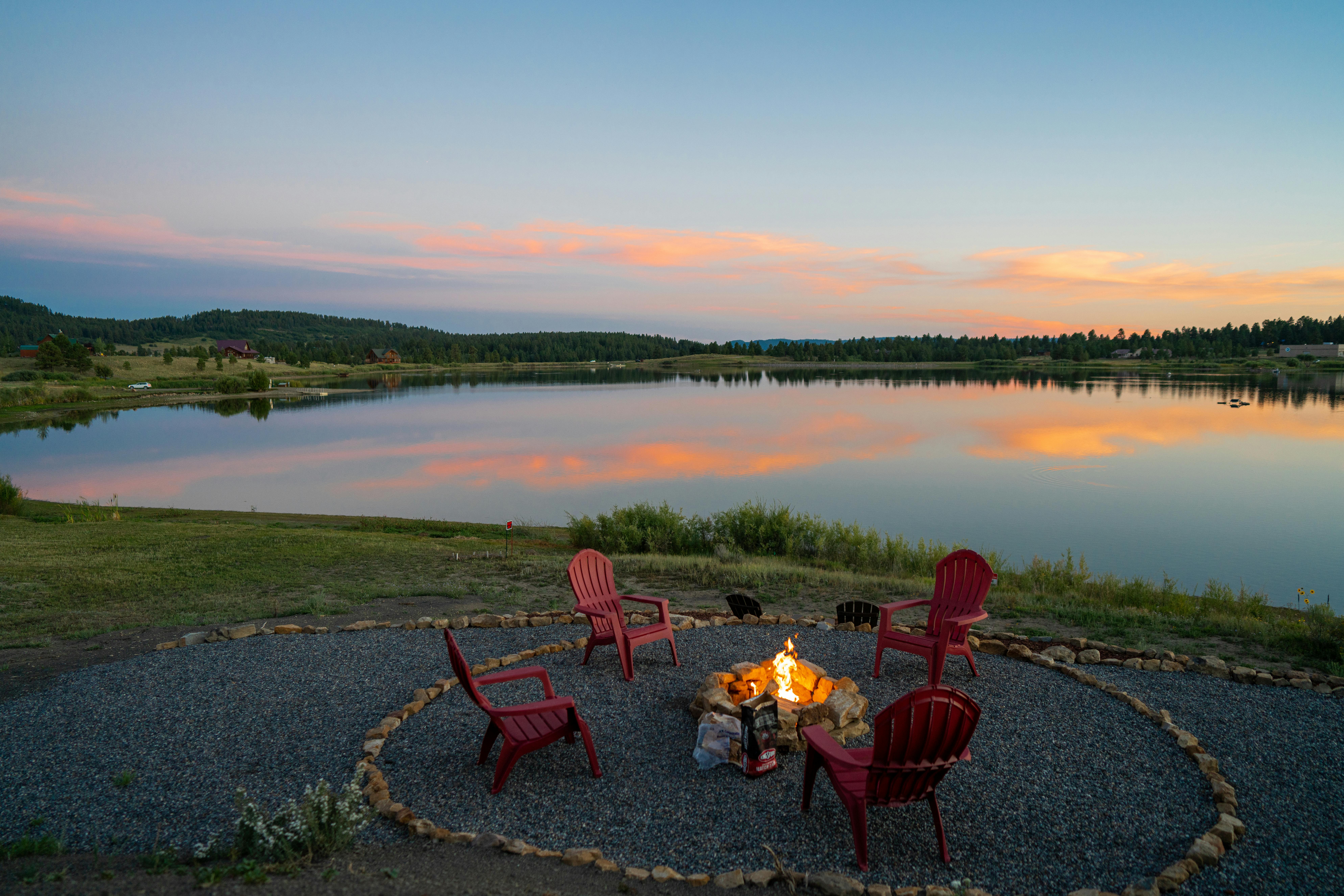 Four red chairs around a campfire beside a lake at sunrise