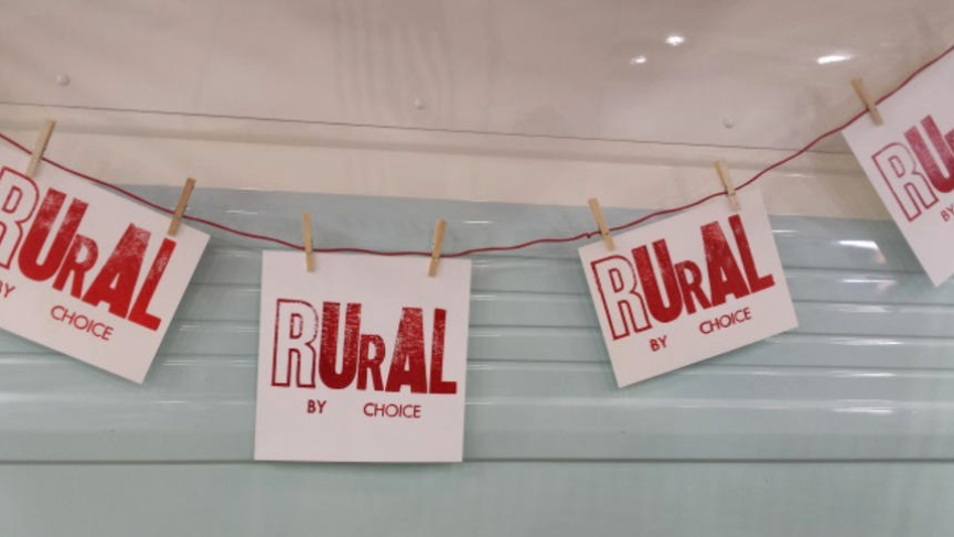 Letterpress printed papers with the words "rural by choice" hang from a string held up by clothespins