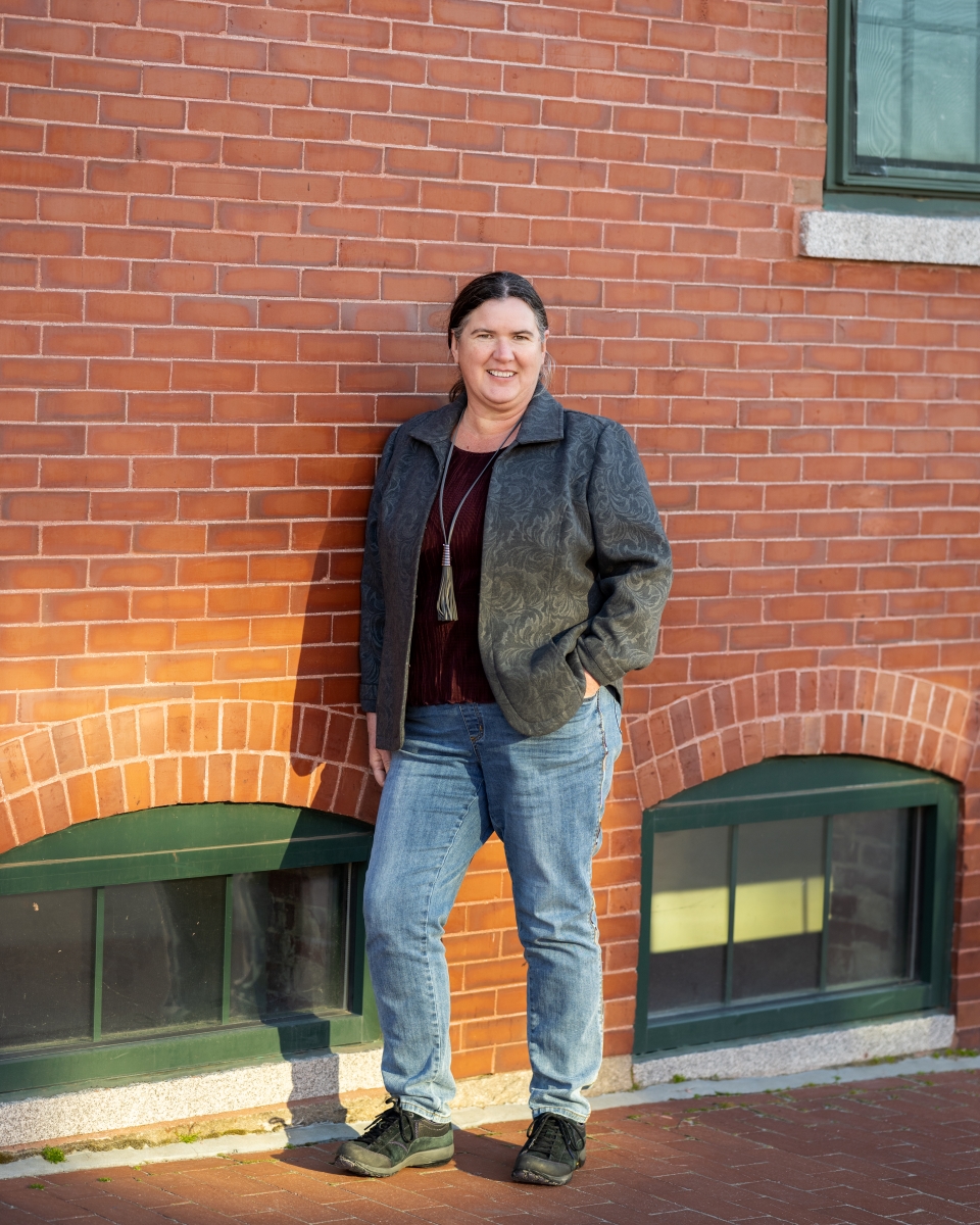 Becky McCray, a light ruddy-skinned woman with dark brown hair, is leaning against a brick wall in a downtown